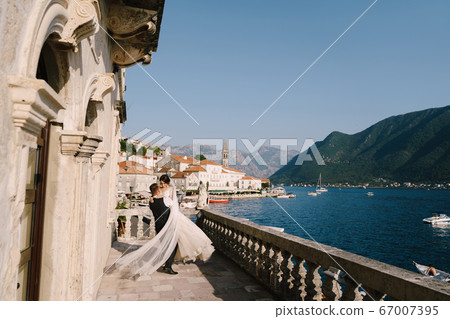 Fine-art wedding photo in Montenegro, Perast. A wedding couple on the hotel terrace with panoramic views of the old town of Perast, the groom circles the bride with a long dress on her hands. 67007395