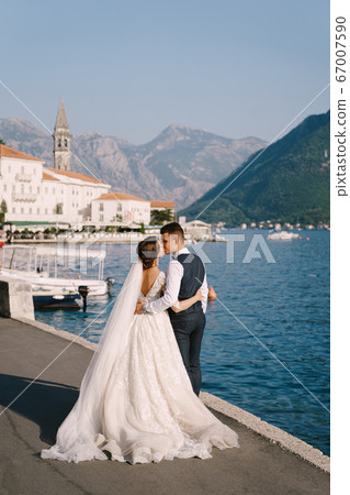 The bride and groom walk along the road and kiss overlooking the city of Perast. Fine-art wedding photo in Montenegro, Perast. The bride and groom walk along the road and kiss overlooking the city of Perast. Fine-art wedding photo in Montenegro, Perast. 67007590