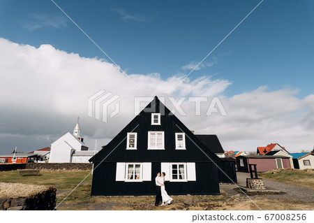 Destination Iceland wedding. Wedding couple near a black wooden house with white windows and shutters. The groom hugs the bride. 67008256