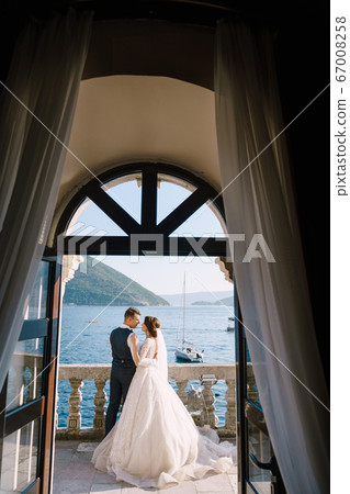 Wedding couple stand on a hotel balcony with sea view, view through an open antique window. Fine-art wedding photo in Montenegro, Perast. Wedding couple stand on a hotel balcony with sea view, view through an open antique window. Fine-art wedding photo in Montenegro, Perast. 67008258