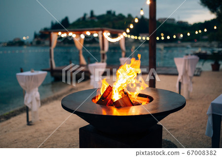 Grill with flames inside. Round table-cooking surface. On the beach, in the background of the gazebo by the water with garlands, in the twilight light. 67008382