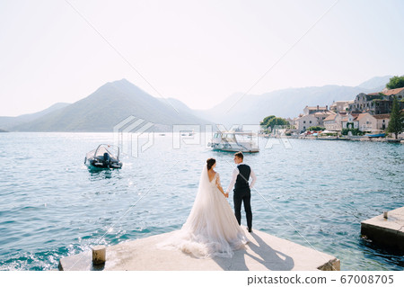 The bride and groom are standing on the pier in the Bay of Kotor, looking at each other, against the background of moored boats, old city and mountains. Fine-art wedding photo in Montenegro, Perast. The bride and groom are standing on the pier in the Bay of Kotor, looking at each other, against the background of moored boats, old city and mountains. Fine-art wedding photo in Montenegro, Perast. 67008705