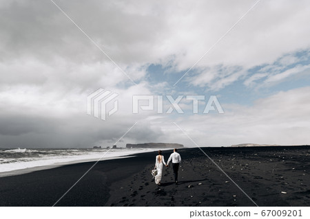 A wedding couple is walking along the black beach of Vic. Sandy beach with black sand on the shores of the Atlantic Ocean. Bride and groom holding hands. Destination Iceland wedding. A wedding couple is walking along the black beach of Vic. Sandy beach with black sand on the shores of the Atlantic Ocean. Bride and groom holding hands. Destination Iceland wedding. 67009201