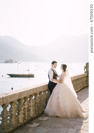 The bride and groom are standing on a large terrace with stone columns, overlooking the Bay of Kotor and the island of St. George and the Island of Gospa od Skrpela at sunset in the old town of Perast 67009330