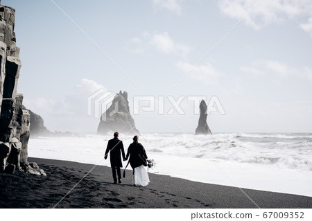 Destination Iceland wedding. A wedding couple walks along the sandy black beach of Vik, near the basalt rock, in the form of pillars. The bride and groom hold hands and walk along the shore. Destination Iceland wedding. A wedding couple walks along the sandy black beach of Vik, near the basalt rock, in the form of pillars. The bride and groom hold hands and walk along the shore. 67009352