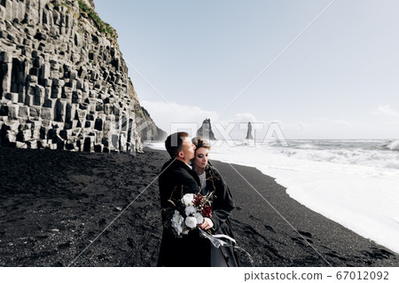 A wedding couple walks along the sandy black beach of Vik, near the basalt rock, in the form of pillars. The bride and groom are hugging on the shore. Destination Iceland wedding. A wedding couple walks along the sandy black beach of Vik, near the basalt rock, in the form of pillars. The bride and groom are hugging on the shore. Destination Iceland wedding. 67012092