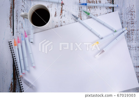 on an wooden table lies an album with many pens and markers and a cup of coffee. 67019303