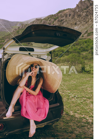 Young beautiful girl in a dress and a wide-brimmed hat near a hatchback car on a background of a mountain landscape. 67024755