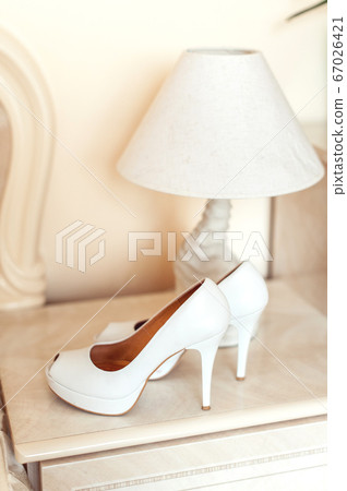 white wedding shoes with heels on a pedestal next to a table lamp. All in bright colors 67026421