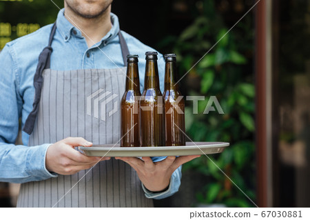 Barman in apron carries tray with bottles of beer in bar 67030881