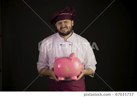 Crying chef man in uniform holds broken pink piggy bank on black background 67035737
