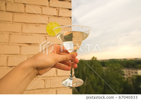 Stay at home balcony cocktail party. Classic home-made Margarita alcohol drink with lime and salt in a hand, trees sky and residential area houses view. Good for website or blog recipes or articles Stay at home balcony cocktail party. Classic home-made Margarita alcohol drink with lime and salt in a hand, trees sky and residential area houses view. Good for website or blog recipes or articles 67040833