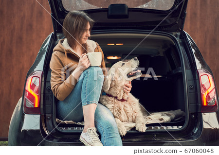 Happy girl with mug in hand with dog sitting in open trunk of black car. 67060048