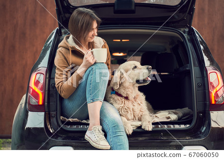 Young girl with mug in hand with dog sitting in open trunk of black car 67060050