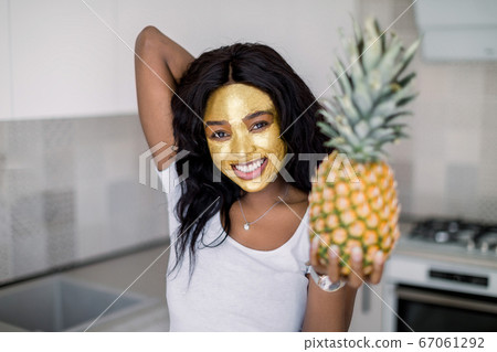 Pretty African woman with golden mask on face, holding fresh pineapple and smiling, posing at camera in light home kitchen interior. Skincare, cleansing, organic cosmetic 67061292