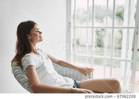 A woman in a T-shirt and shorts sits in a chair near the window A woman in a T-shirt and shorts sits in a chair near the window 67062208