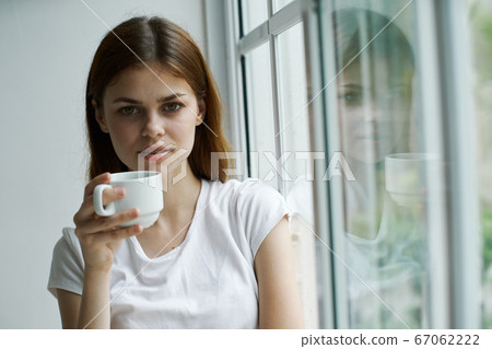 calm woman with a cup near the window looks at the camera  67062222
