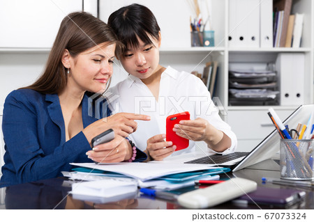 Two women write sms by phone in the office Two women write sms by phone in the office 67073324