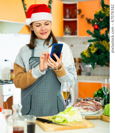 Woman in xmas hat using phone during cooking christmas dinner Woman in xmas hat using phone during cooking christmas dinner 67076382