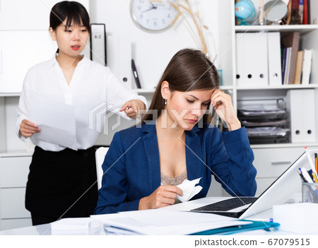Frustrated woman sitting at office desk with disgruntled boss behind 67079515