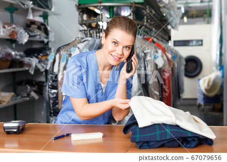 Smiling girl worker of laundry talking on phone while checking clothes taken for dry cleaning Smiling girl worker of laundry talking on phone while checking clothes taken for dry cleaning 67079656