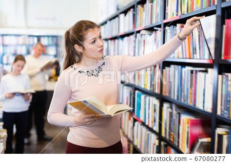 Young attractive female looking for necessary books on shelves in bookstore 67080277