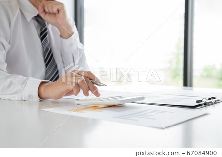 A businessman is sitting at a desk and calculating financial graphs about real estate investment expenditures A businessman is sitting at a desk and calculating financial graphs about real estate investment expenditures 67084900