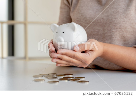 A young woman who holds a piggy bank and puts a coin on the saving display table expands the growing business for success and saving for retirement A young woman who holds a piggy bank and puts a coin on the saving display table expands the growing business for success and saving for retirement 67085152