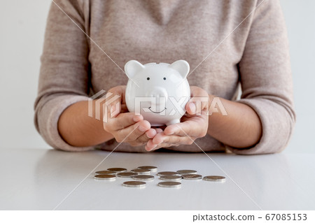 A young woman who holds a piggy bank and puts a coin on the saving display table expands the growing business for success and saving for retirement A young woman who holds a piggy bank and puts a coin on the saving display table expands the growing business for success and saving for retirement 67085153