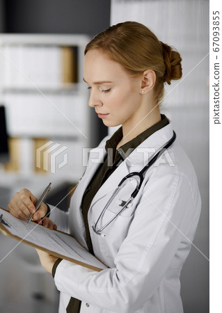 Cheerful smiling female doctor using clipboard in clinic. Portrait of friendly physician woman at work. Perfect medical service in hospital. Medicine concept Cheerful smiling female doctor using clipboard in clinic. Portrait of friendly physician woman at work. Perfect medical service in hospital. Medicine concept 67093855