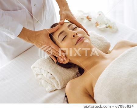 Beautiful brunette woman enjoying facial massage with closed eyes. Relaxing treatment in medicine and spa center concepts Beautiful brunette woman enjoying facial massage with closed eyes. Relaxing treatment in medicine and spa center concepts 67093924