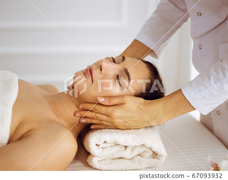 Beautiful brunette woman enjoying facial massage with closed eyes. Relaxing treatment in medicine and spa center concepts 67093932