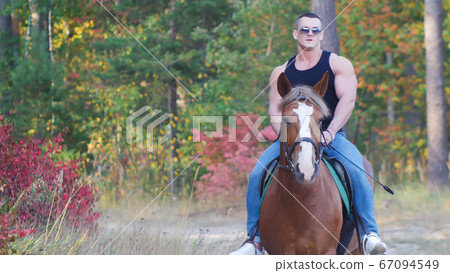 A strong man with a powerful body in a black t-shirt in sunglasses rides a horse A strong man with a powerful body in a black t-shirt in sunglasses rides a horse 67094549