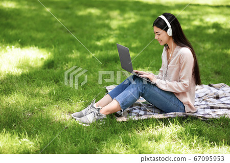 Smiling asian girl relaxing with laptop and headphones on lawn in park Smiling asian girl relaxing with laptop and headphones on lawn in park 67095953