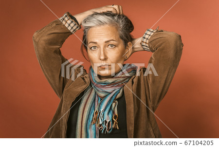 Pensive gray-haired woman makes hairstyle, collecting hair with his hands on the back of head while posing in studio on orange background. Tinted image 67104205