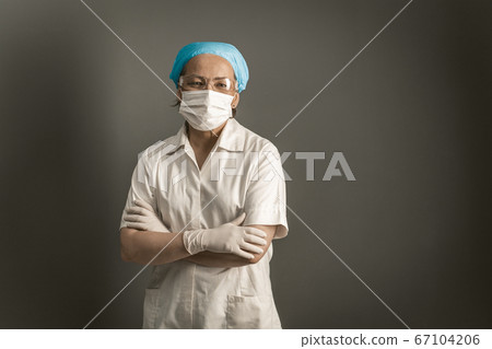 Doctor crossed her arms looking at camera. Female caucasian medic in protective mask and white uniform stands on gray background. Empty space for text at right. Tinted image 67104206