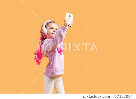 Pretty modern girl with pink dreadlocks in headphones smiling and making selfie on smartphone isolated over yellow 67132094
