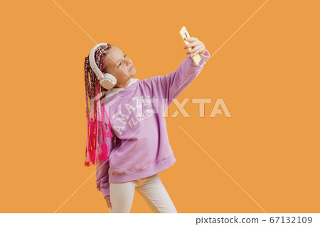 Pretty modern girl with pink dreadlocks in headphones smiling and making selfie on smartphone isolated over yellow 67132109