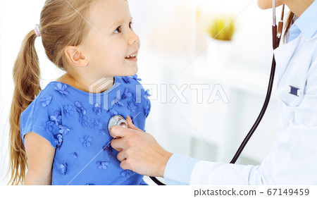 Doctor examining a child by stethoscope in sunny clinic. Happy smiling girl patient dressed in blue dress is at usual medical inspection Doctor examining a child by stethoscope in sunny clinic. Happy smiling girl patient dressed in blue dress is at usual medical inspection 67149459