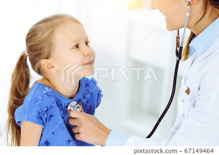 Doctor examining a child by stethoscope in sunny clinic. Happy smiling girl patient dressed in blue dress is at usual medical inspection 67149464