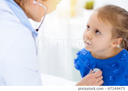 Doctor examining a child by stethoscope in sunny clinic. Happy smiling girl patient dressed in blue dress is at usual medical inspection Doctor examining a child by stethoscope in sunny clinic. Happy smiling girl patient dressed in blue dress is at usual medical inspection 67149473