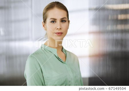 Beautiful adult business woman dressed in green blouse standing straight in sunny office. Business headshot Beautiful adult business woman dressed in green blouse standing straight in sunny office. Business headshot 67149504