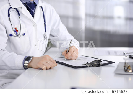 Unknown male doctor sitting and working with clipboard of medication history record in clinic at his working place, close-up. Young physician at work. Perfect medical service, medicine concept 67156283