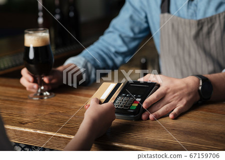 Bar business. Barman in apron gives glass of beer and terminal, girl pays bill with card Bar business. Barman in apron gives glass of beer and terminal, girl pays bill with card 67159706