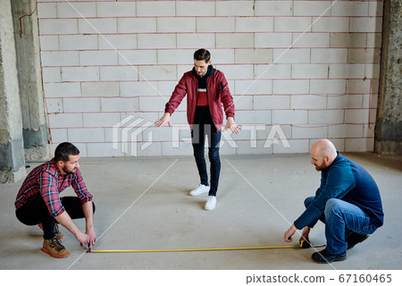 Two young builders sitting on squats while holding ends of measuring tape 67160465