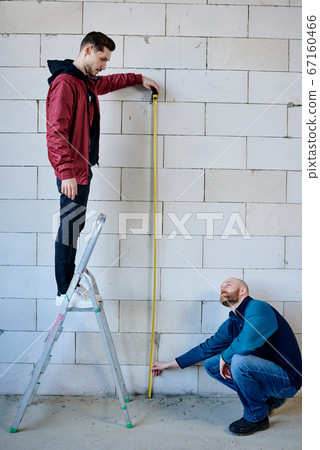 Young builder standing on stepladder and holding measuring tape by brick wall 67160466