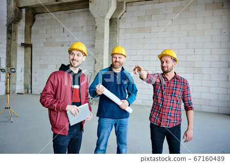 Happy young engineer in hardhat pointing at one of walls of unfinished building 67160489