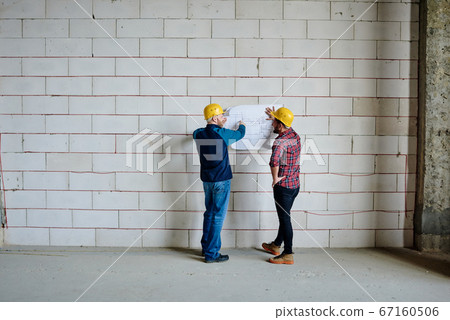 Two professional engineers in hardhats standing by wall and discussing sketch Two professional engineers in hardhats standing by wall and discussing sketch 67160506