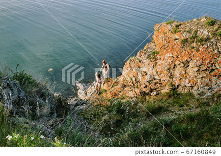 Two young backpackers in casualwear moving down stone rock along waterside 67160849