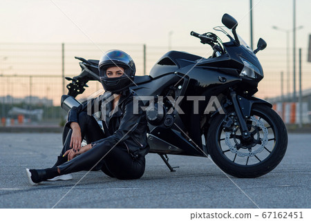 Biker woman in black leather jacket and full face helmet sits near stylish sports motorcycle at urban parking. Traveling and active lifestyle concept. 67162451